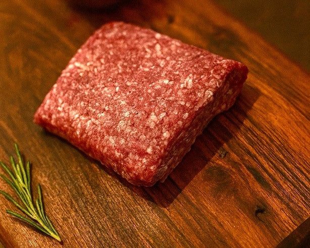 Raw ground beef log on wooden cutting board with small bowl of seasoning