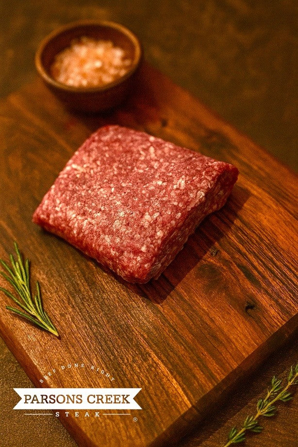 Raw ground beef log on wooden cutting board with small bowl of seasoning