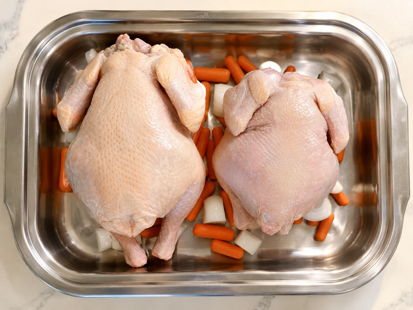 Two raw whole chickens with skin, arranged in a metal tray with carrots.